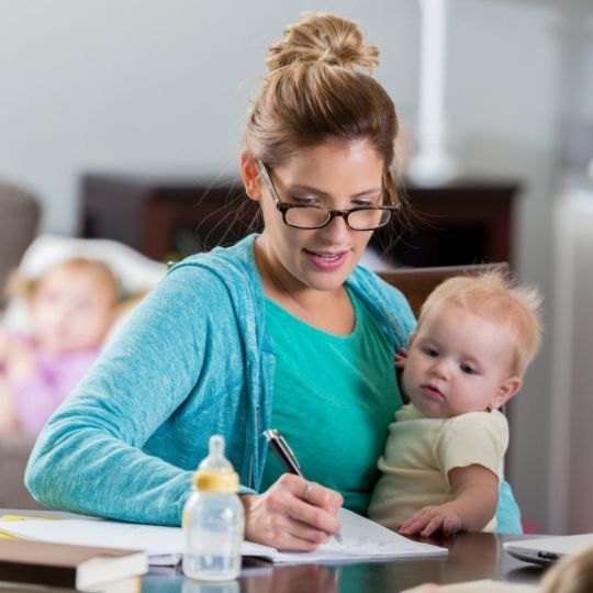 woman sitting at table, writing a note while holding a baby